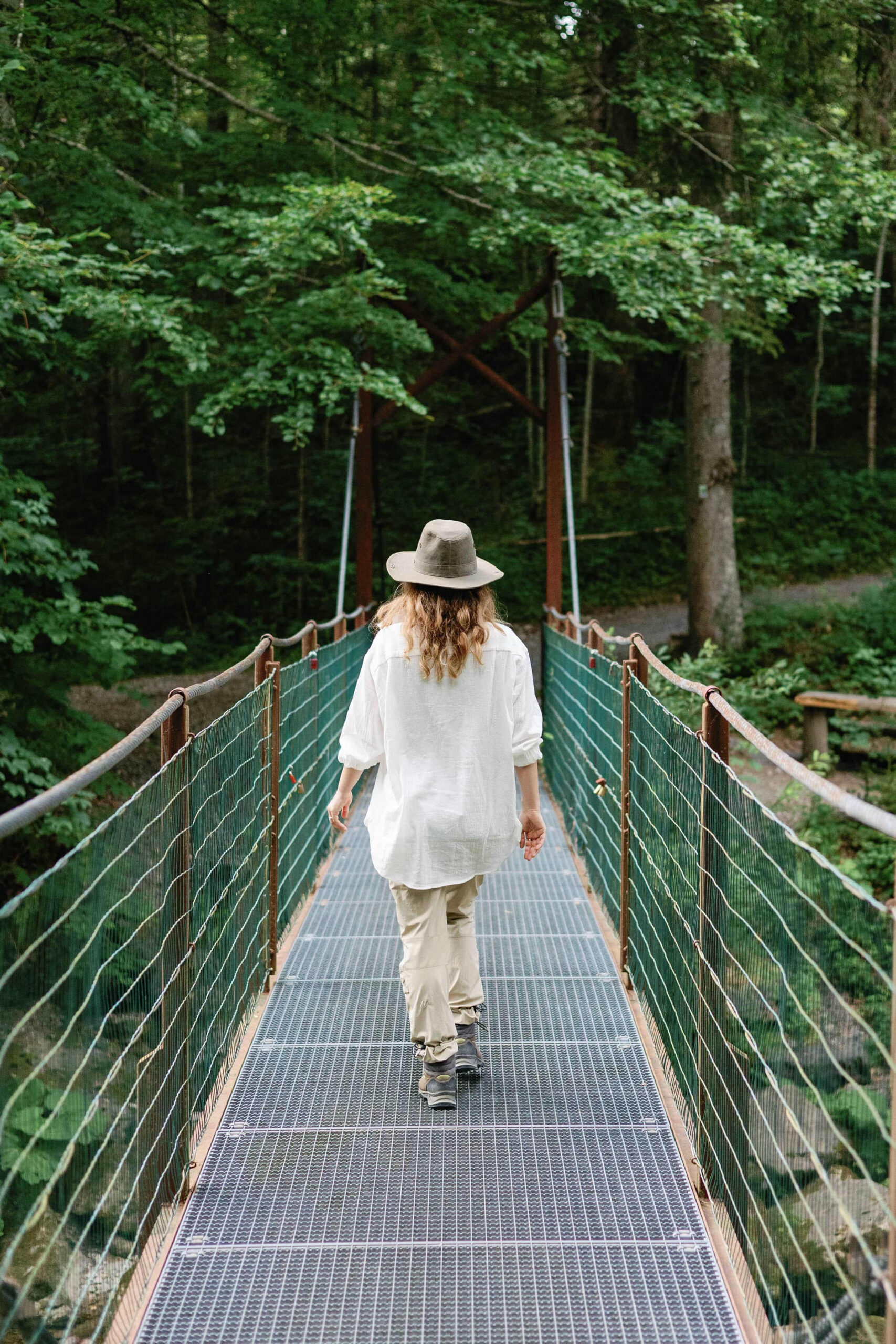 Woman slowing down while walking across a suspension bridge surrounded by green trees.